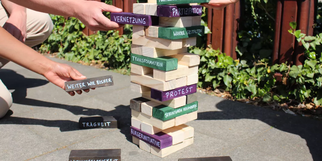 Hände, die einen Jenga-Turm bauen, dessen Steine mit Schlüsselwörtern aus der Bildungsarbeit versehen sind